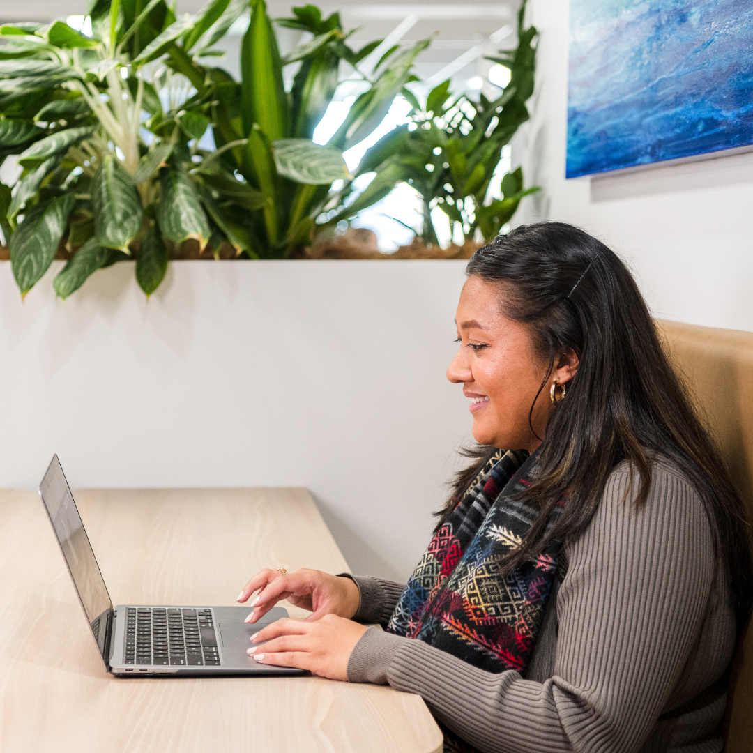 Woman typing on a laptop at a table, smiling. Plants are behind her.