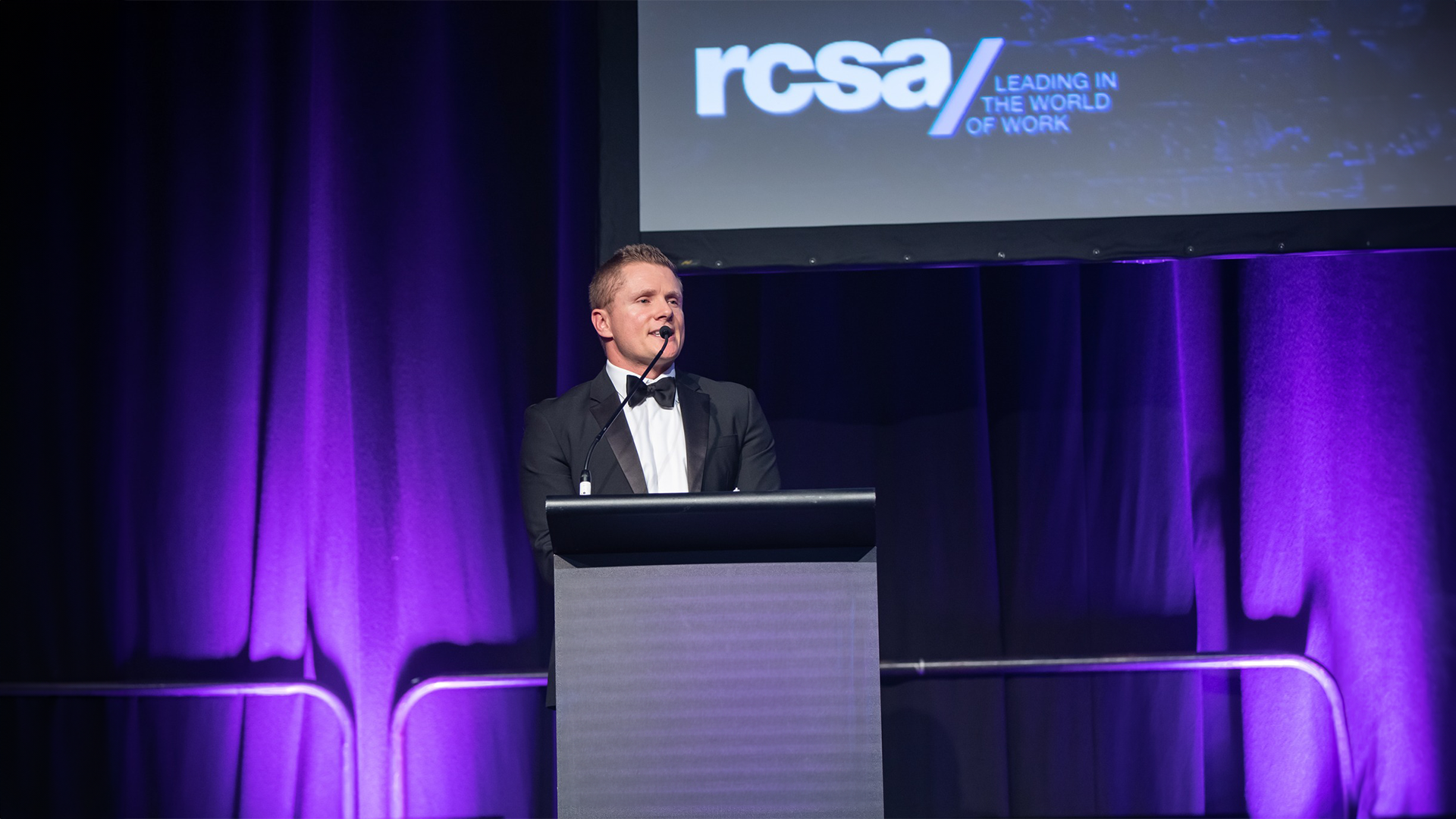Man in tuxedo speaks at podium with 