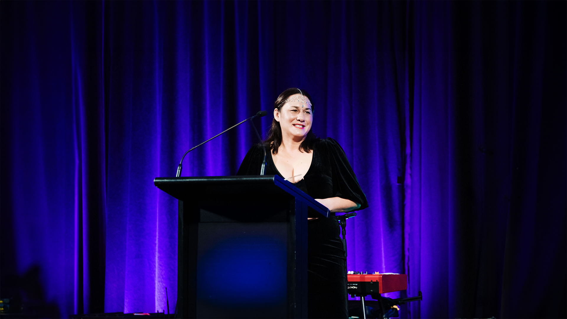 Woman speaking at a podium on stage with blue curtains.