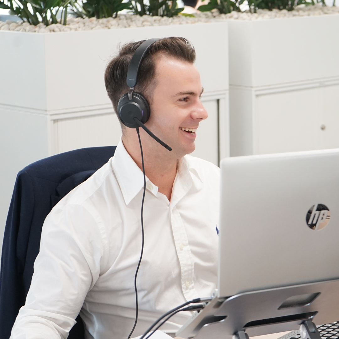 Man wearing headset smiling, working on laptop in an office setting.