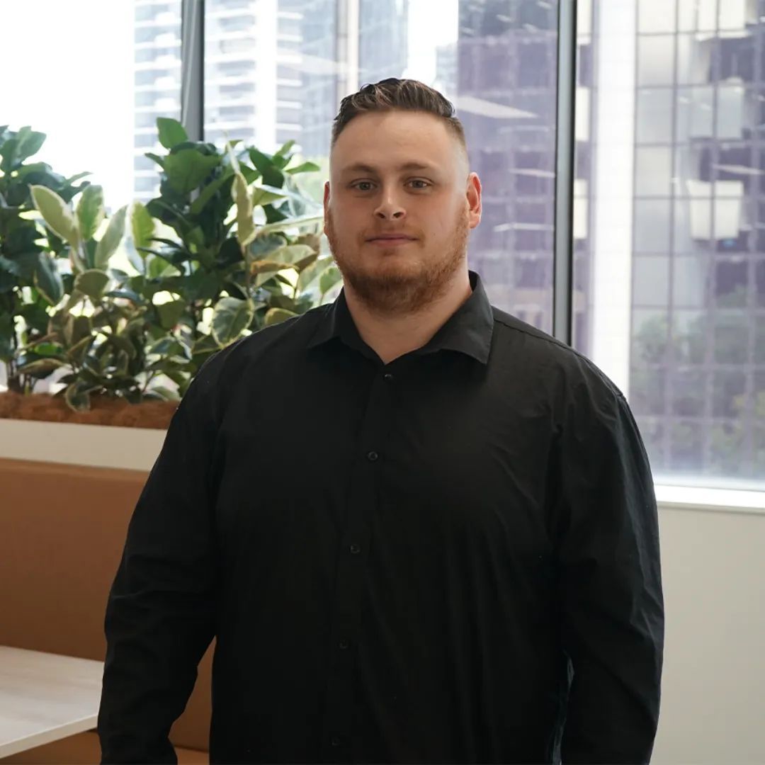Man in a black shirt, standing near a window with greenery and a city view.