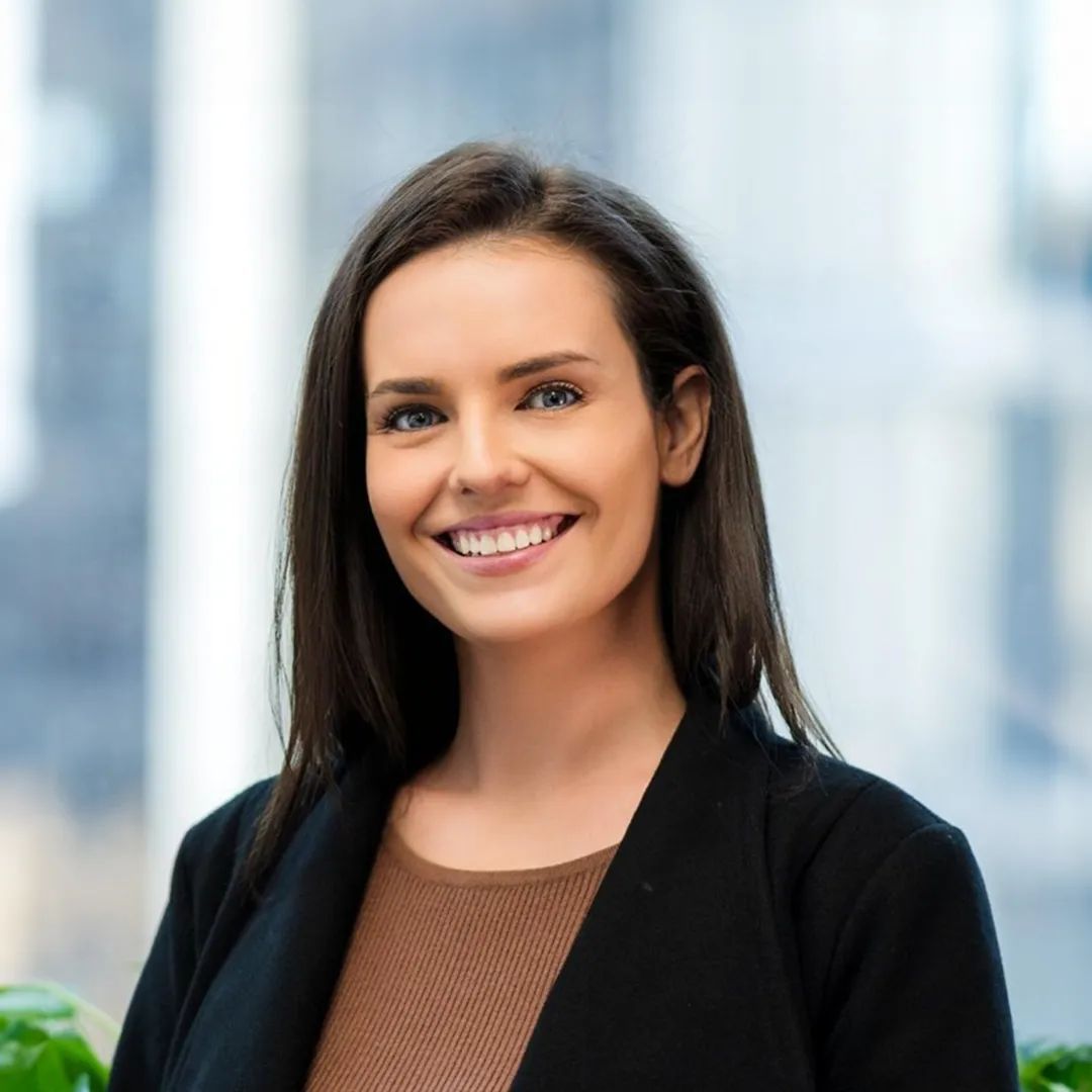 Woman smiling, wearing a black blazer and brown top, indoors with a blurred background.