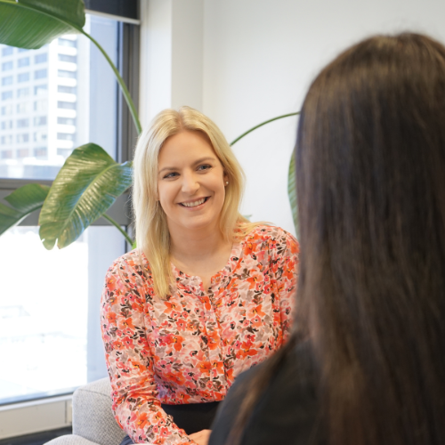 Woman in blue blazer, smiling, talking on a phone, near a window with plant.