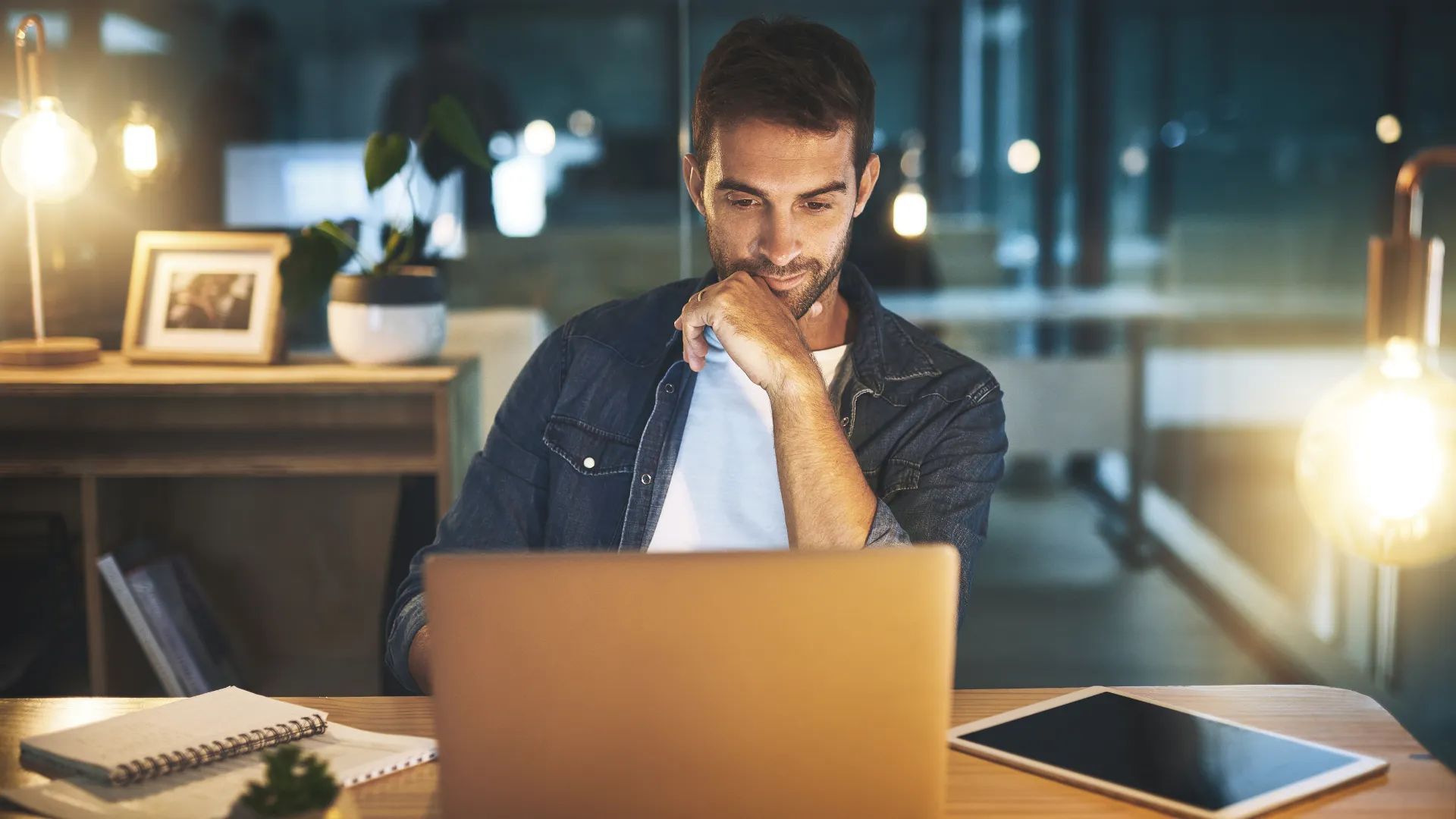 Man working intently on laptop at desk, lit by lamps in a dimly lit room.