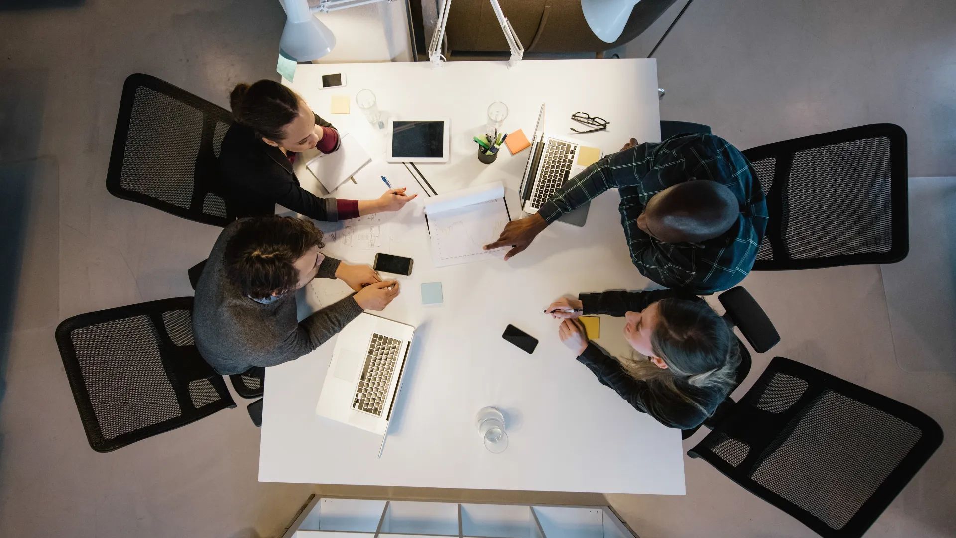 People sitting around a white table, reviewing documents and using laptops in an office setting.