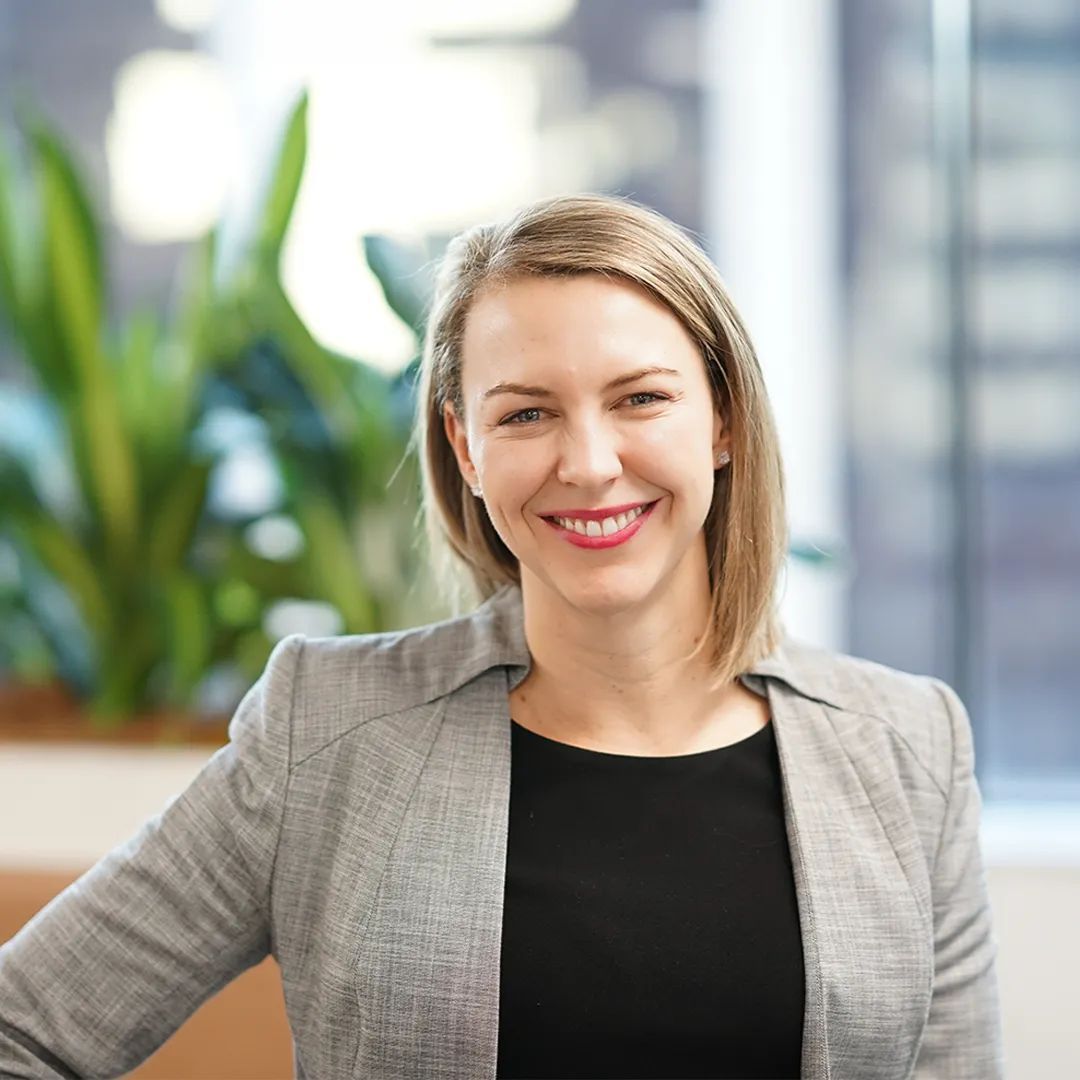 Woman smiling, wearing a gray blazer and black top. Indoor office setting with green plants.