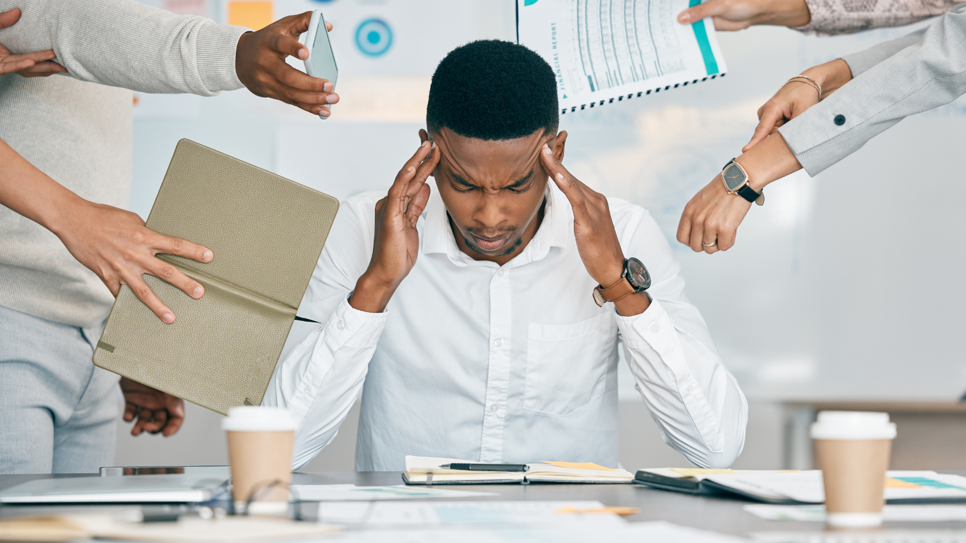 Person with hands on head surrounded by hands holding papers, stressed at desk.