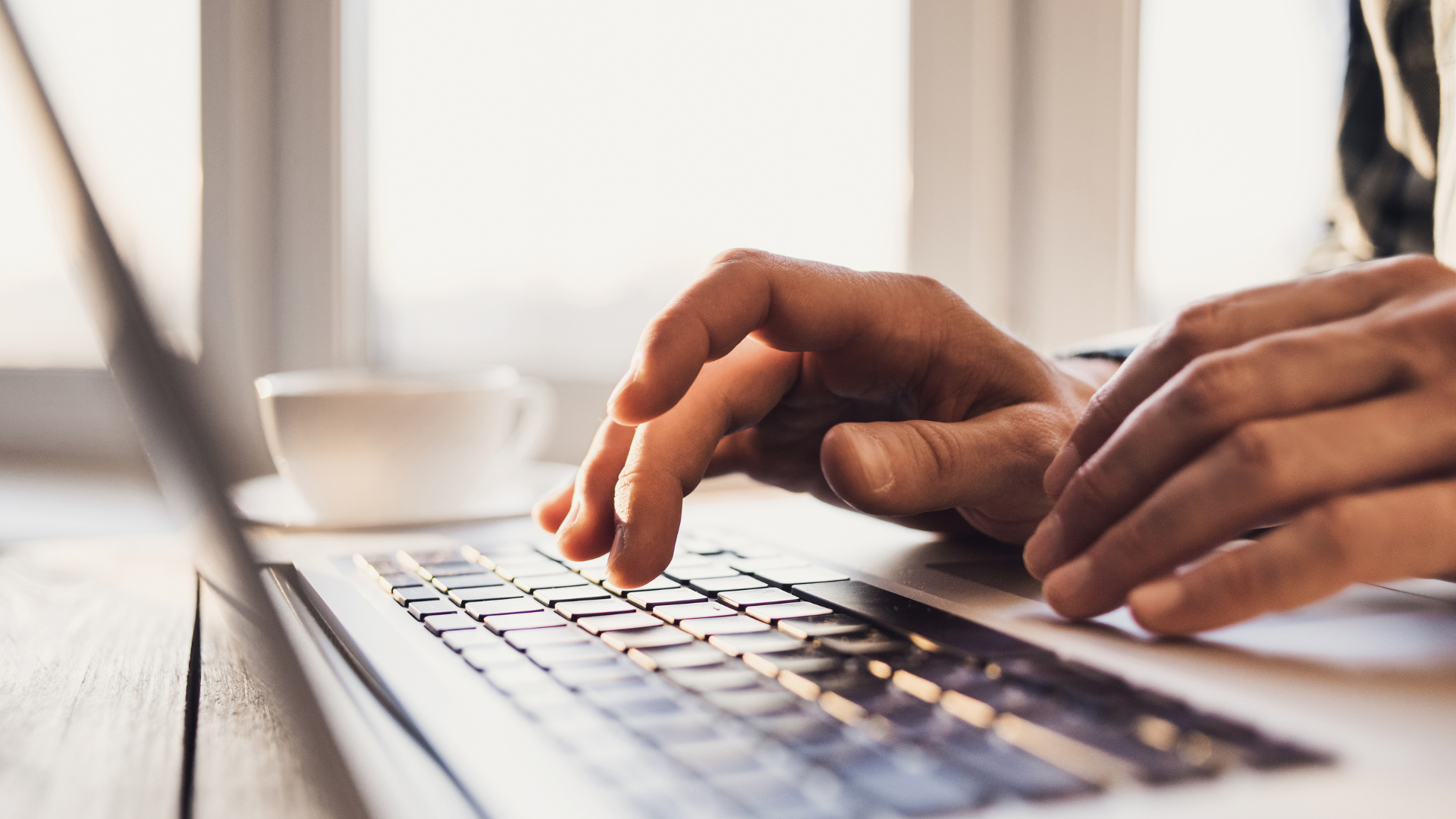 Hands typing on a laptop keyboard; a cup of coffee is visible in the background by a window.