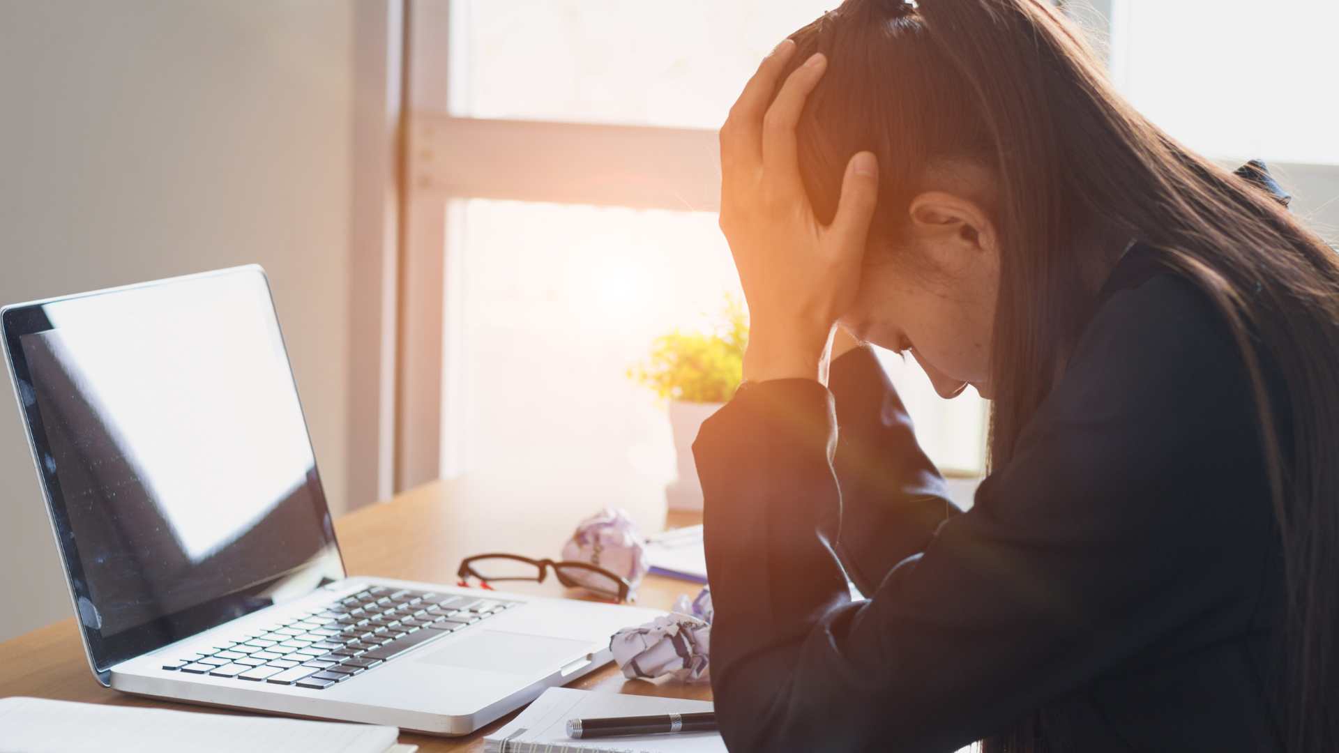 Woman with hands on head, looking down at laptop, stressed at desk.