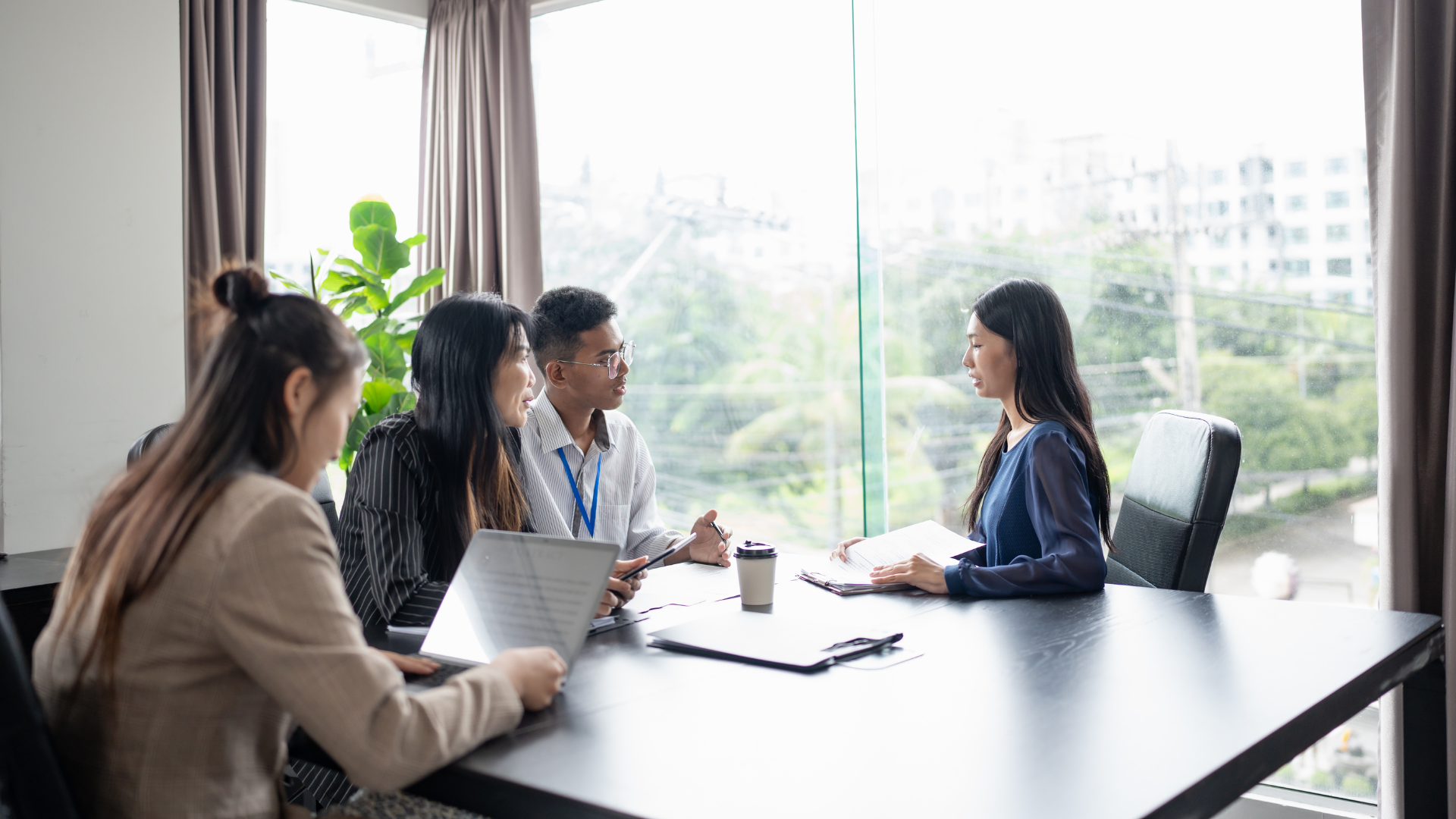 Four people in meeting at a table, discussing documents. Large window and plants in the background.