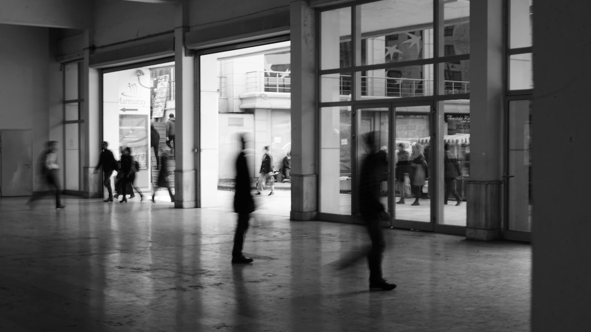 Black and white photo of a large open space with blurred figures walking. Sunlight streams in from open doors and windows.