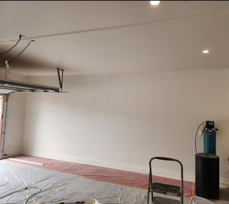 Garage interior with a painted white ceiling and walls, covered floor, ladder, and equipment.