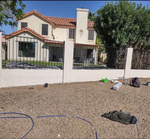 Beige two-story house with red tile roof, white wall, black fence in front yard with gravel and scattered items.