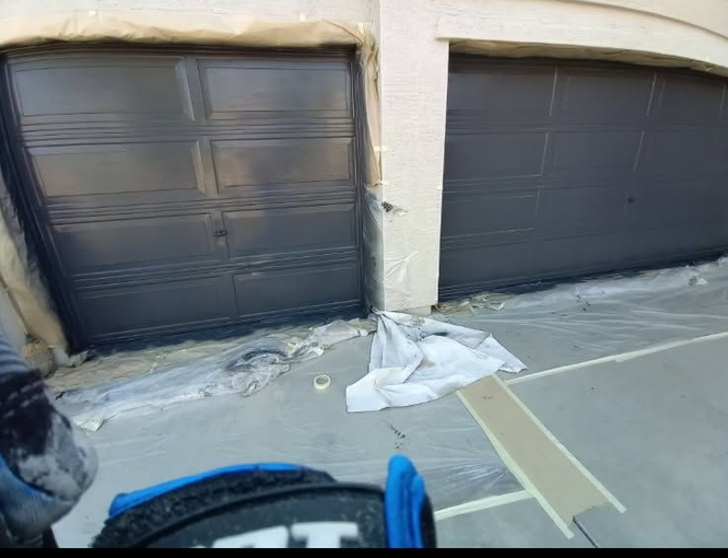 Two garage doors painted dark gray with painter's tape and plastic for protection.