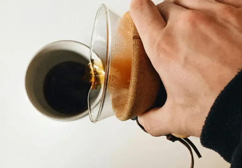 Hand pouring coffee from a cup into a glass mug with a brown sleeve