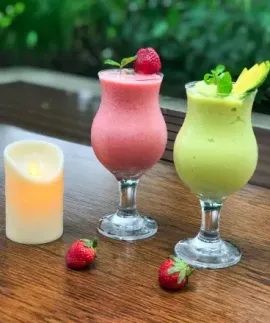 Two colorful cocktails on a wooden table with a lit candle and raspberries, outdoors.