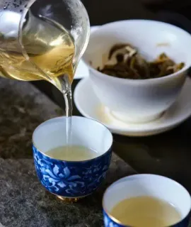 Tea being poured into a blue patterned cup beside another cup and a white bowl of tea leaves.
