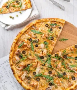 Overhead view of a vegetable pizza on a wooden board with a slice removed, beside a plate and utensils