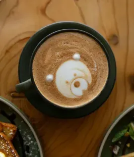 Top-down view of a latte in a green mug on a wooden table, with food plates partly visible nearby