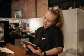 Barista at a café counter taking an order on a tablet while holding a phone to her ear