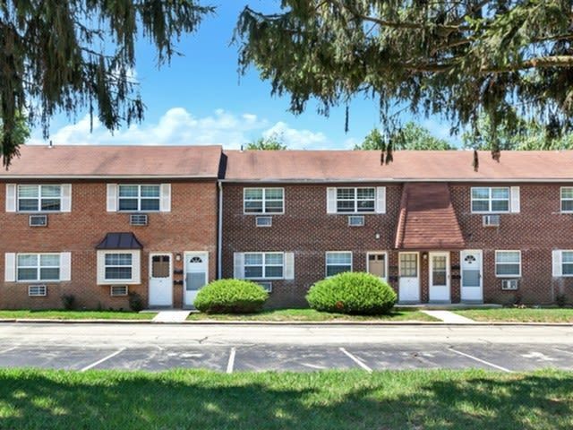 Row of brick apartment buildings with front doors, windows, and trimmed shrubs along a parking lot.