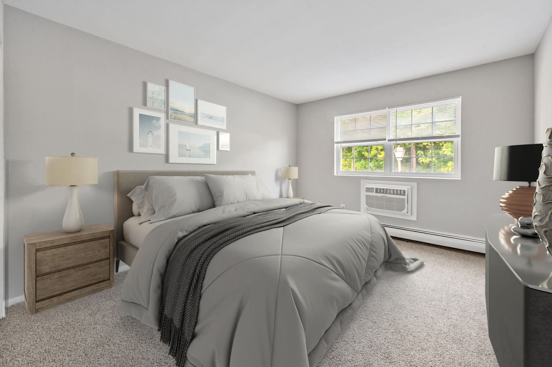 Bedroom in an apartment with a made bed, two nightstands, lamps, wall art, and a window AC unit.