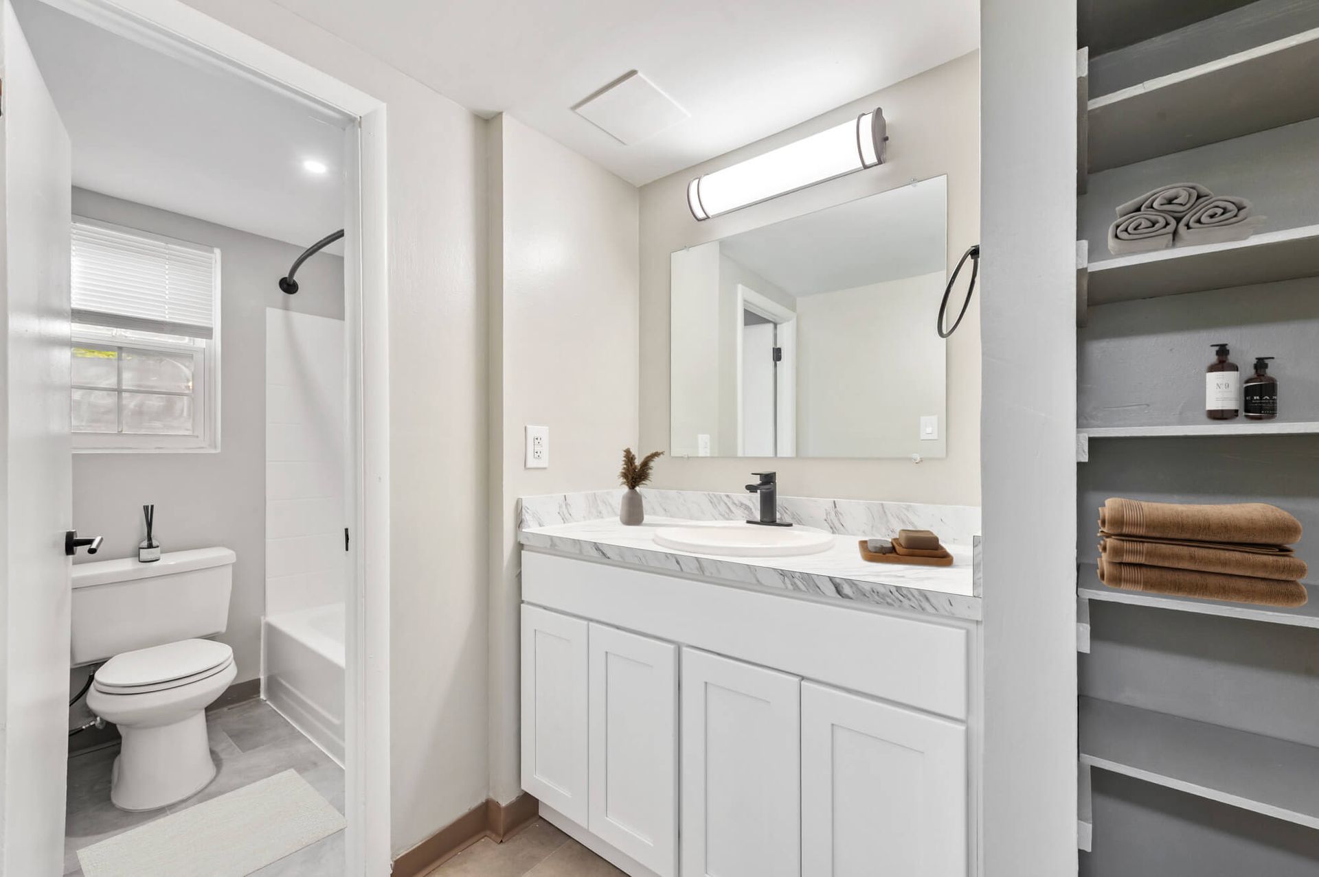 Bright bathroom with white vanity, marble countertop, large mirror, and shelves holding towels.