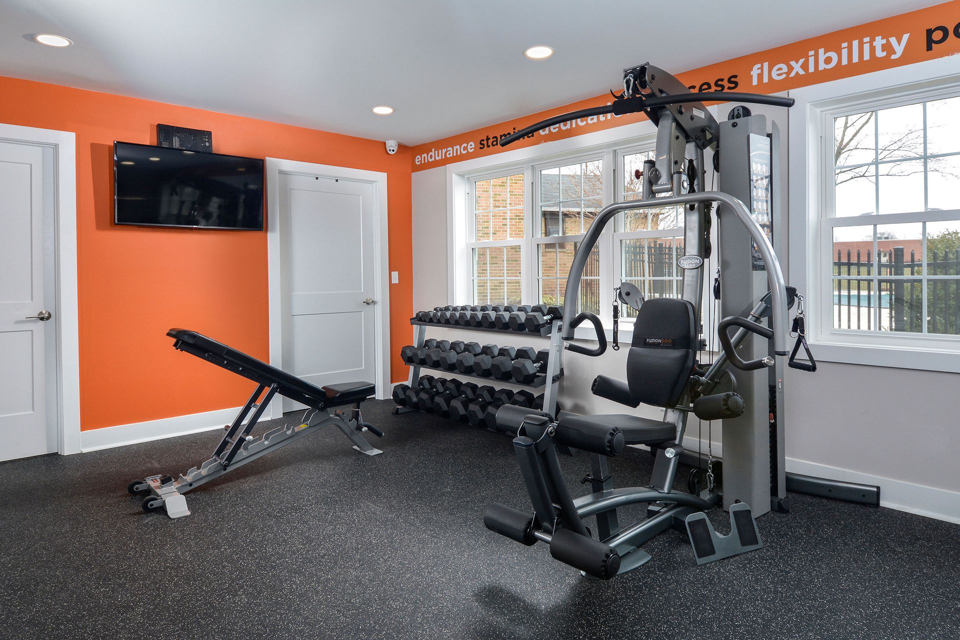 Bright community gym with orange wall, dumbbell rack, bench, and a multi-station machine by windows.