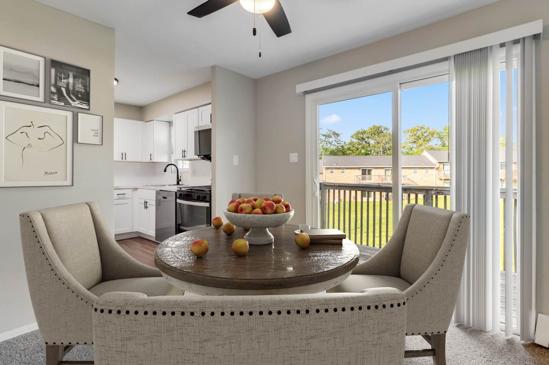Dining area with round wooden table and beige upholstered chairs; open kitchen in the background; sliding balcony door.