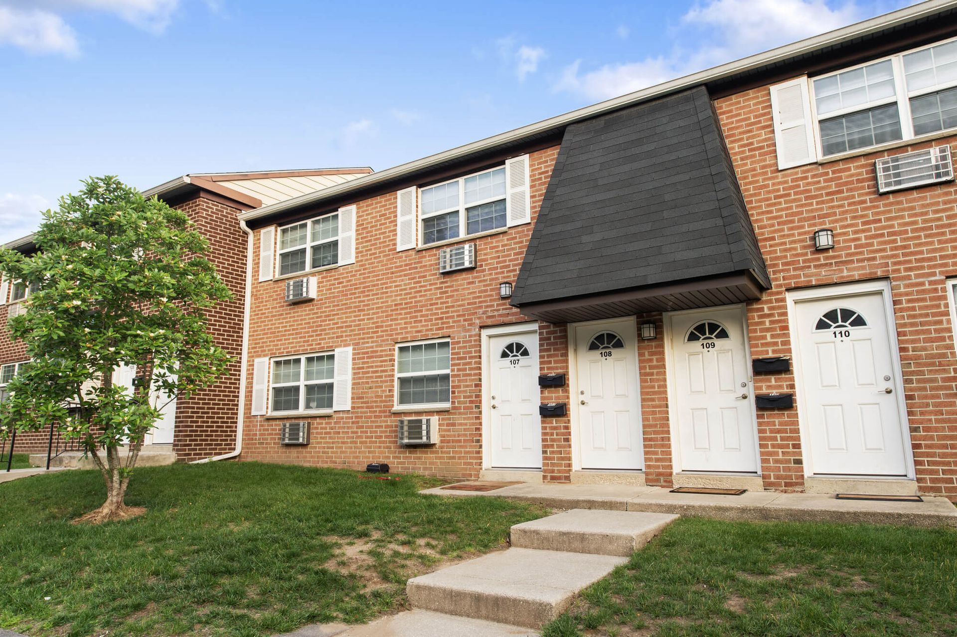 Exterior view of a brick apartment building with white doors and a small tree in front.