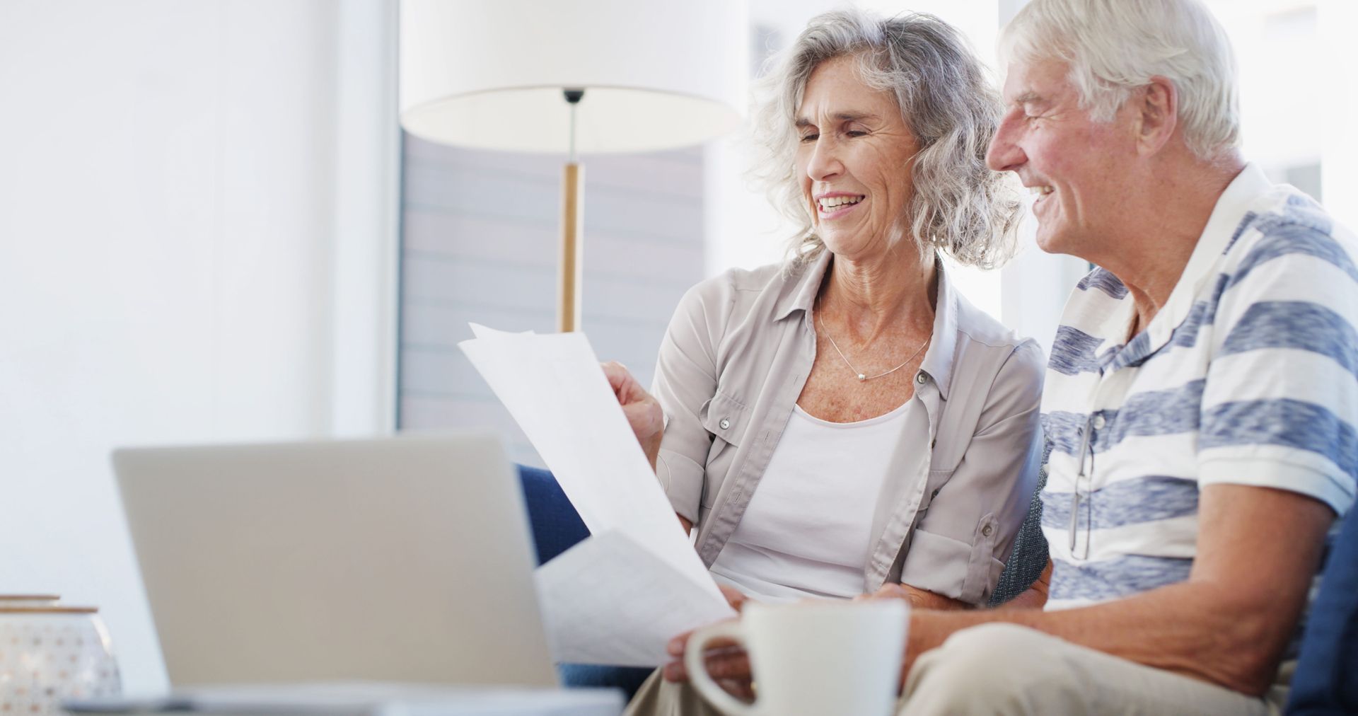 Two seniors smiling and reviewing documents beside a laptop in a bright living room