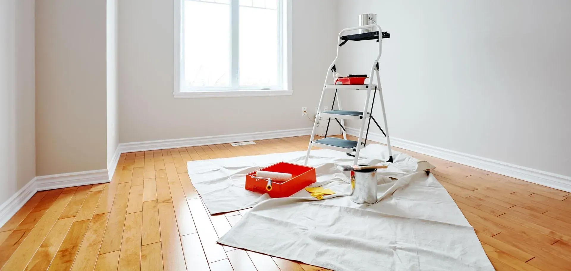 Ein Raum mit hellgrauen Wänden und Holzboden, in dem eine Trittleiter und eine rote Farbschale befinden.