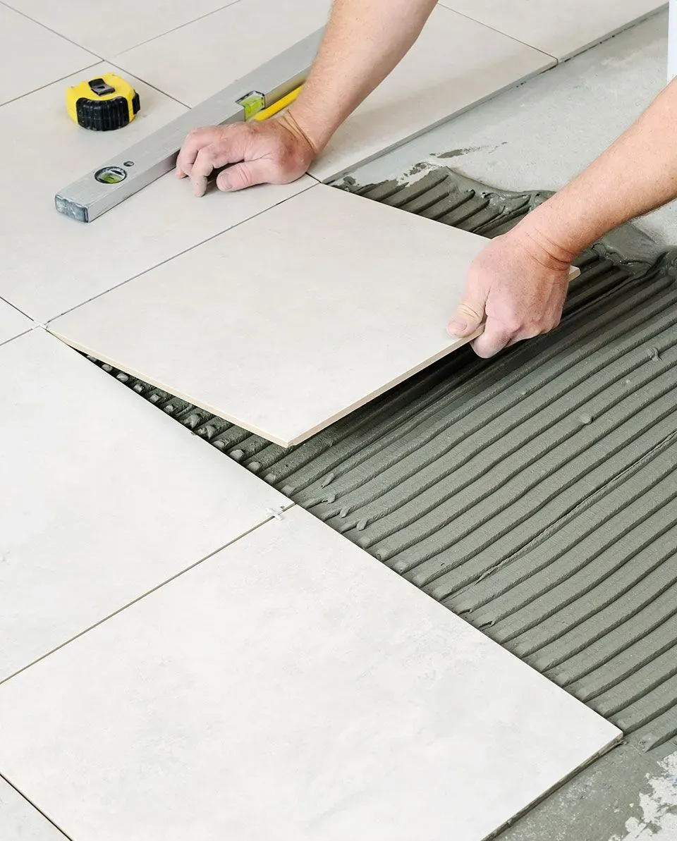Eine Person verlegt quadratische weiße Fliesen auf grauen Mörtelrillen auf einem Boden;