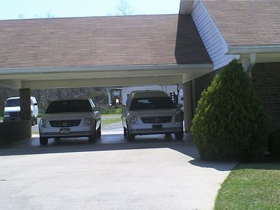 Two silver SUVs parked under a carport with a brown roof. A small evergreen bush is on the right.