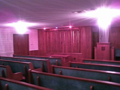 Interior of a chapel with rows of pews, a wooden stage, and bright pink lighting.
