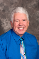 Man with white hair, smiling widely, wearing a blue shirt and patterned tie. Gray background.
