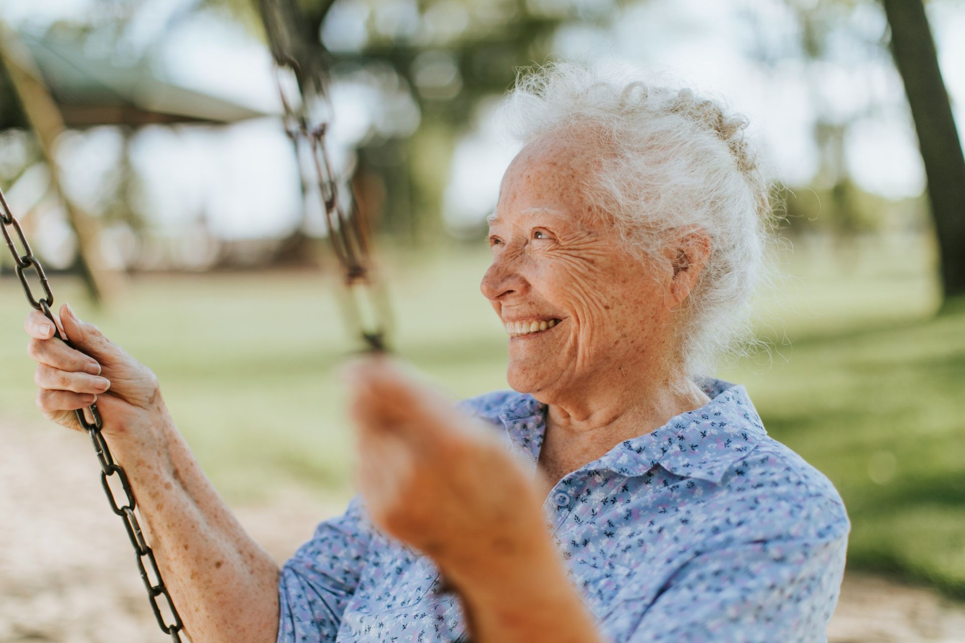 An elderly woman is sitting on a swing in a park and smiling.