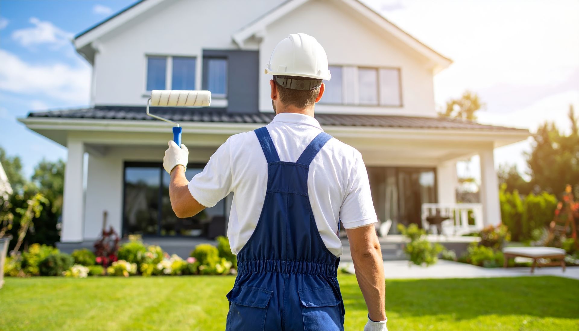 Painter in blue overalls holding a paint roller in front of a house.