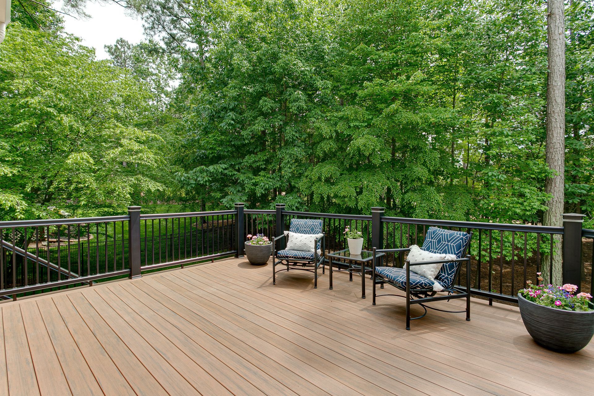 A wooden deck with two patterned armchairs, a small side table, and two potted plants overlooking a dense green forest.