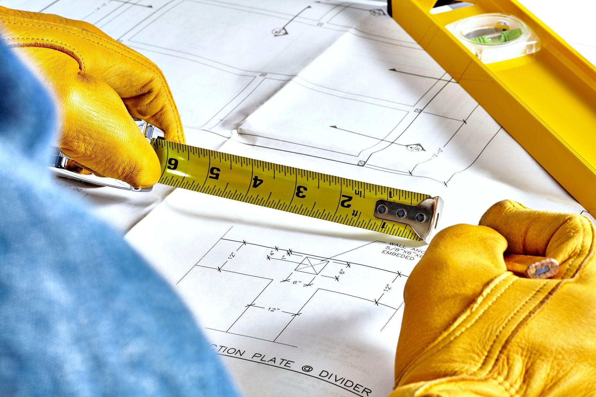 Hands in yellow work gloves measure a blueprint with a yellow tape measure, with a level visible in the background.