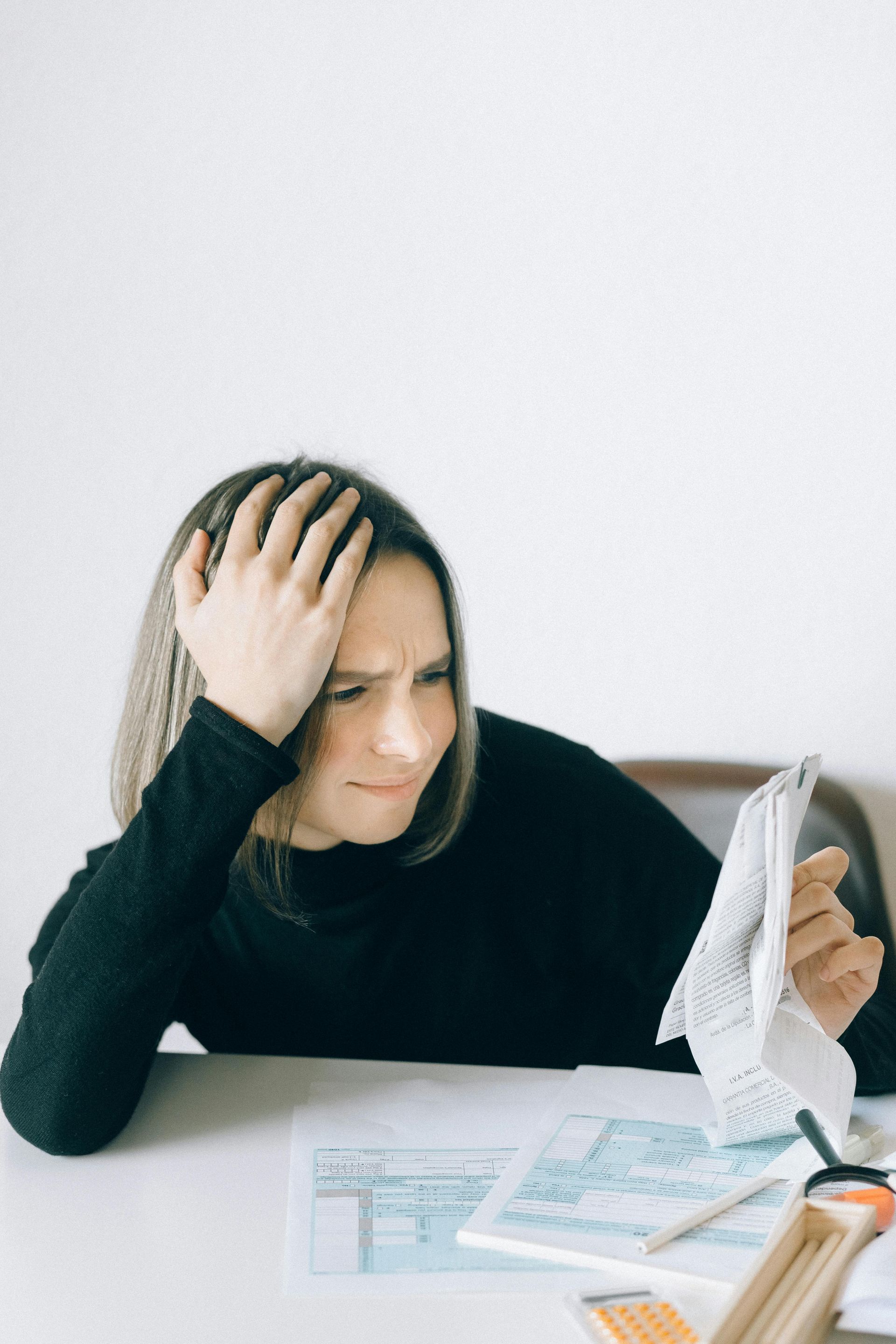 A woman is sitting at a table holding her head and looking at a piece of paper.