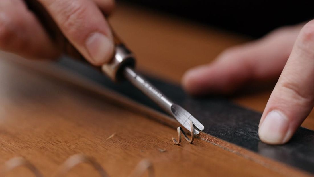 Manos utilizando una herramienta de tallado para dar forma a la madera a lo largo de un borde recto.
