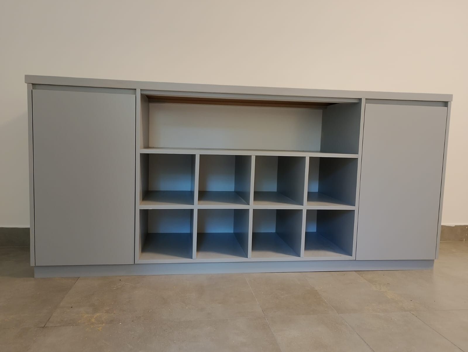 Gabinete de almacenamiento gris con estantes abiertos y puertas corredizas, contra una pared blanca y un piso color canela.