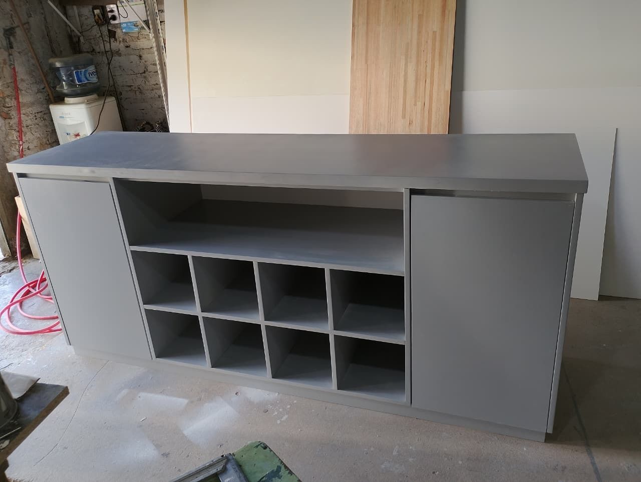 Mueble gris con estantes centrales abiertos para botellas y dos armarios laterales.