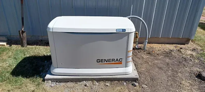 A gray and white Generac home generator is on a concrete pad near a building, with a gas line connected to it.