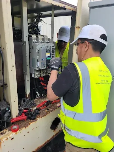 Two electricians working on electrical equipment, wearing safety vests and hard hats.