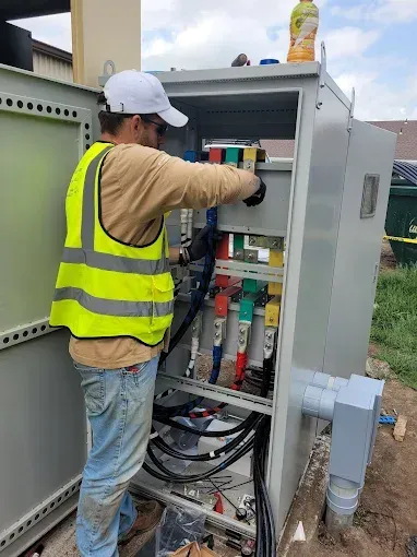 Electrician in a safety vest working on a grey electrical panel outdoors, wires visible.