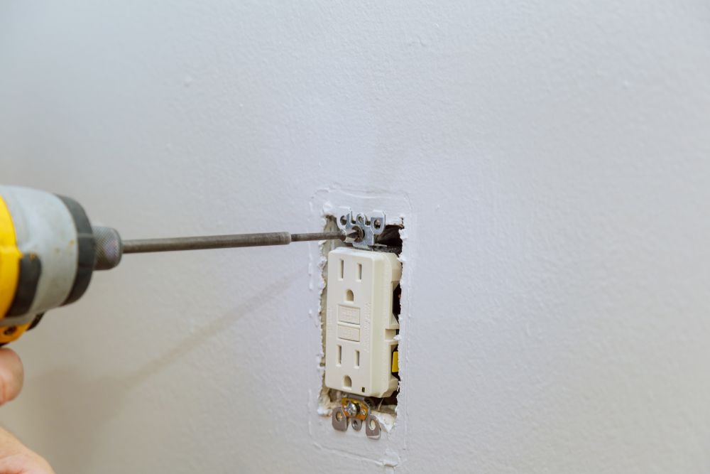 Person using a power drill to install an electrical outlet in a white wall.