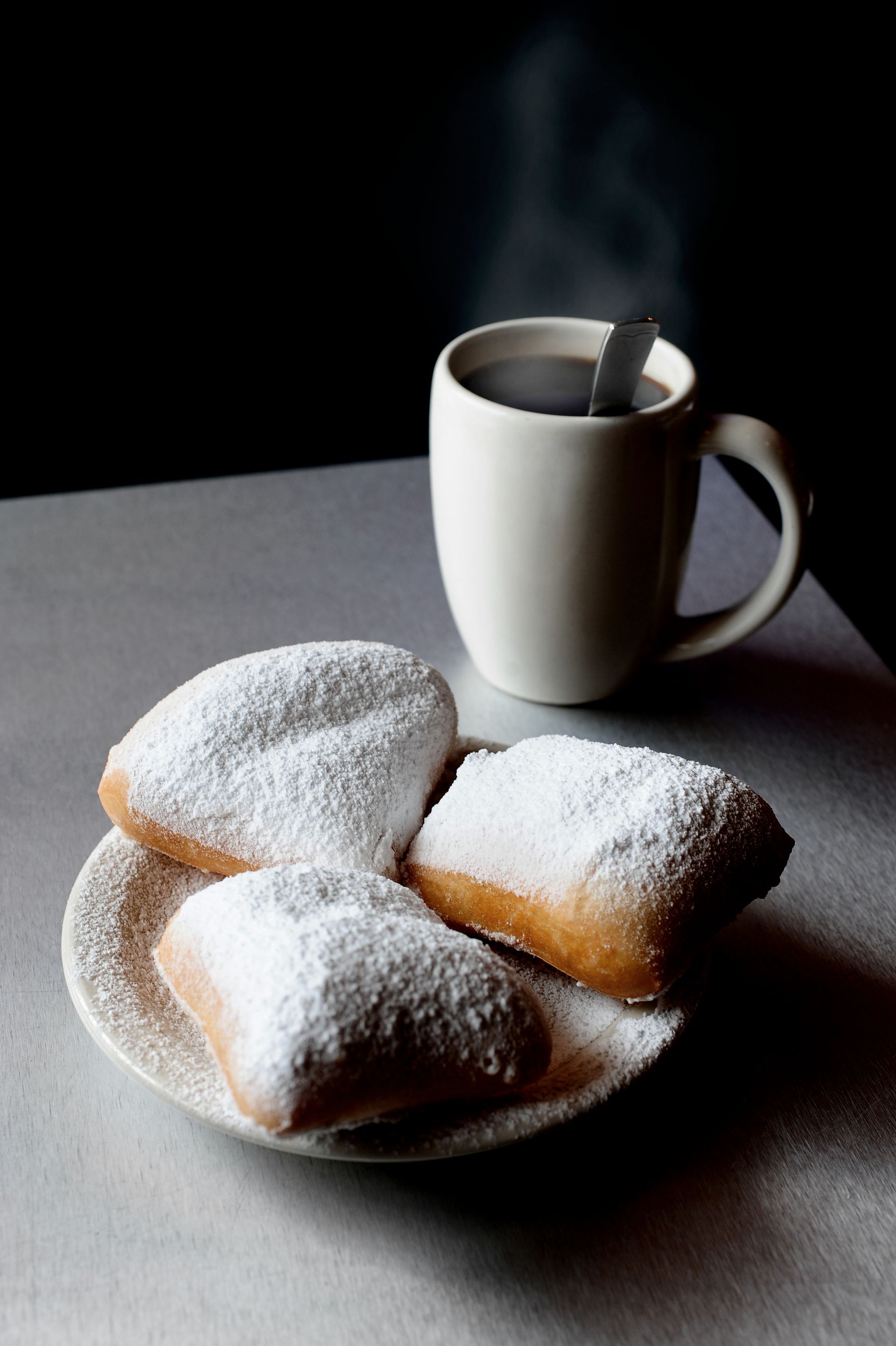 A plate of powdered sugar donuts next to a cup of coffee