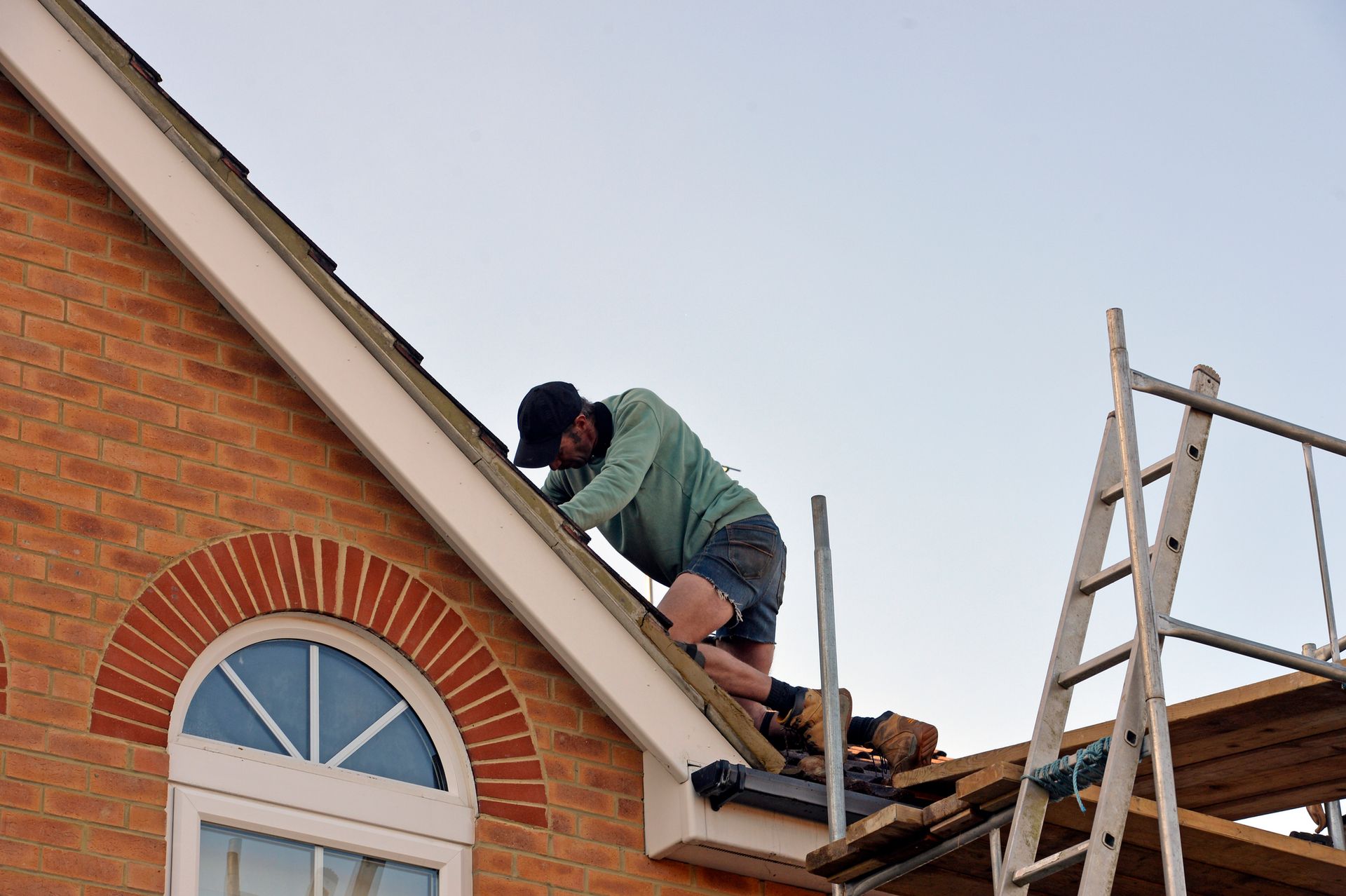 Worker repairing brick house roof using ladder and scaffolding.